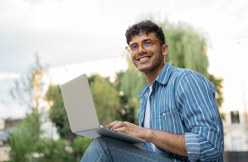 Ein junger Student sitzt lächelnd mit seinem Laptop im Freien und freut sich über beglaubigte Übersetzungen zu einem attraktiven Studentenpreis.